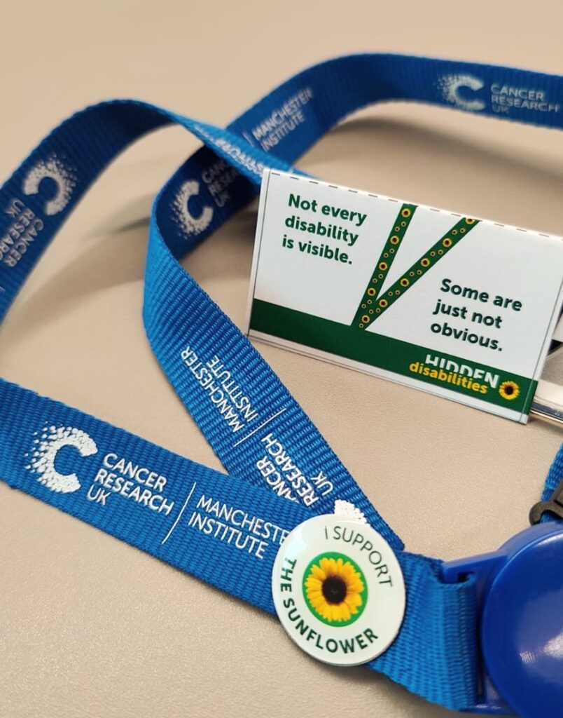 A Cancer Research UK Manchester Institute lanyard laid on a table showing the Hidden Disabilities supporters badge pinned to it