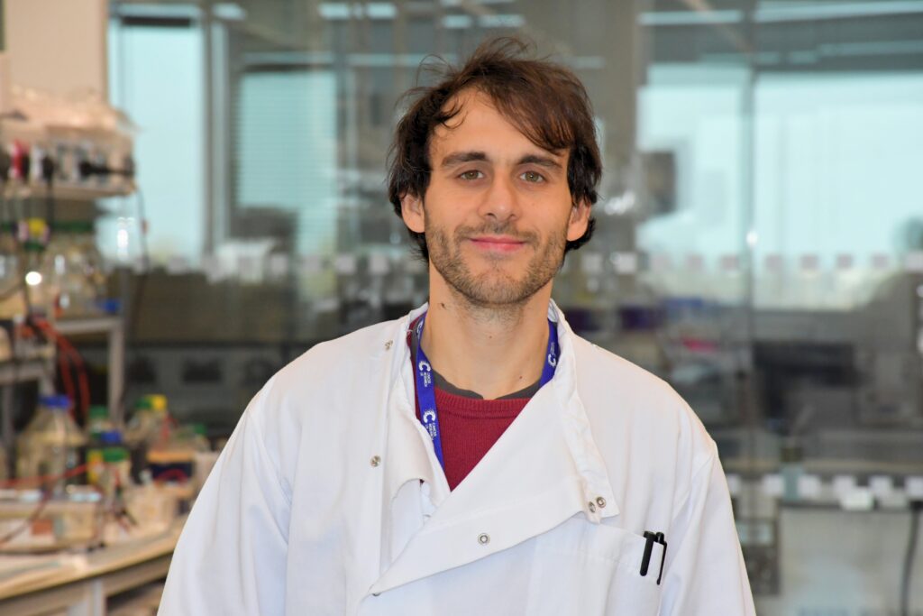 Photograph of Dr Luciano Nicosia wearing a lab coat standing in front of a lab window